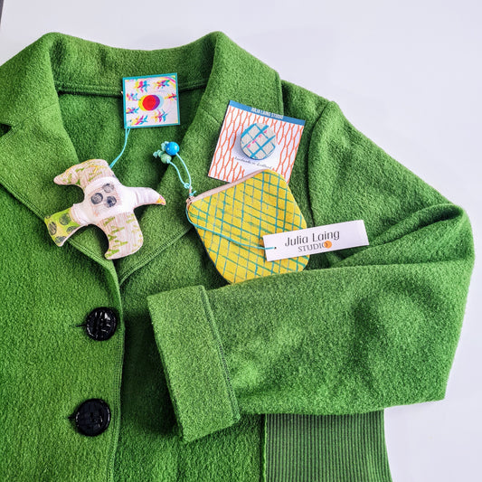A handmade textile hanging bird, a yellow purse and a hand printed linen brooch are displayed on a bright green wool jacket, which is lying flat on top of a white table.