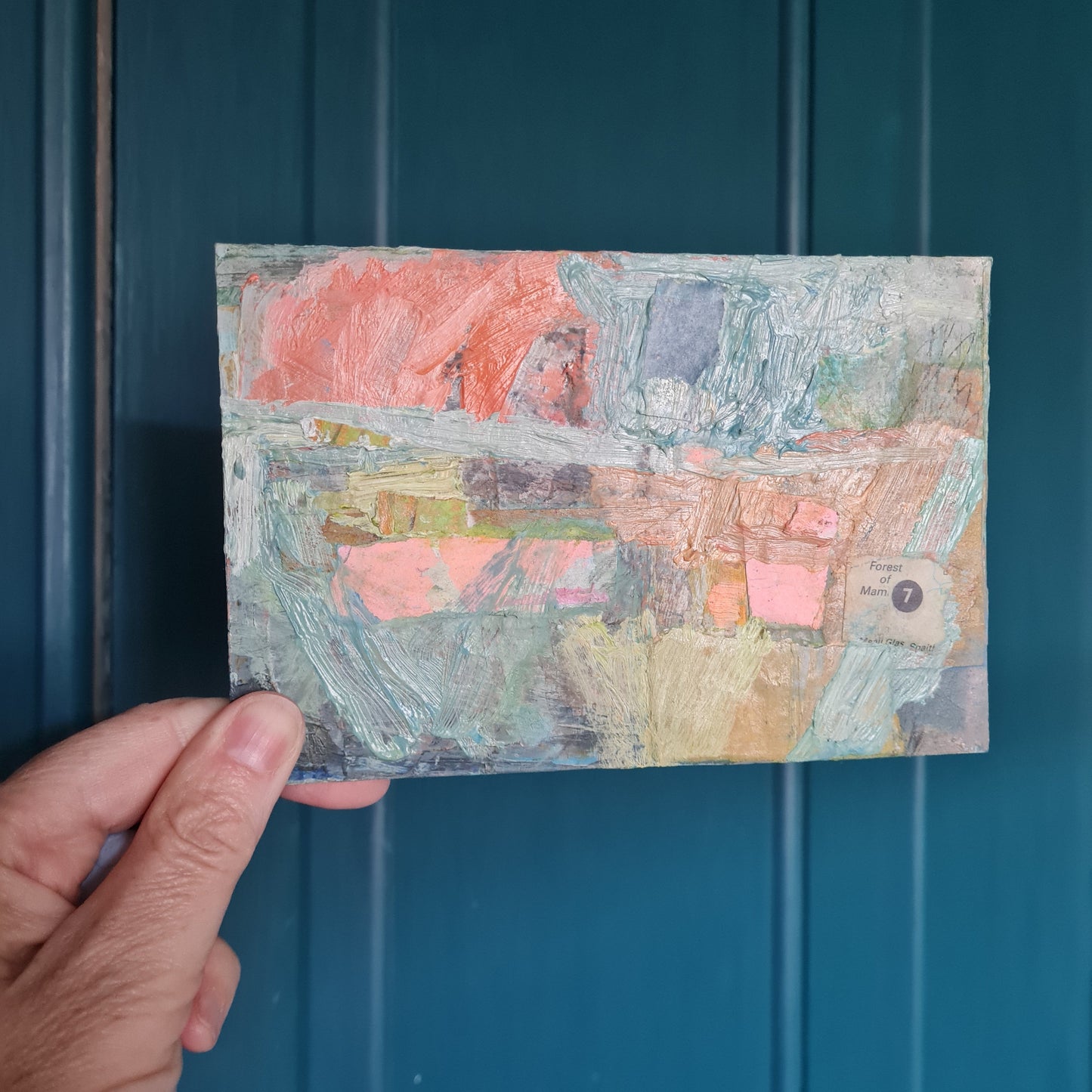 Hand holding a textured, multicolored postcard size painting against a teal background