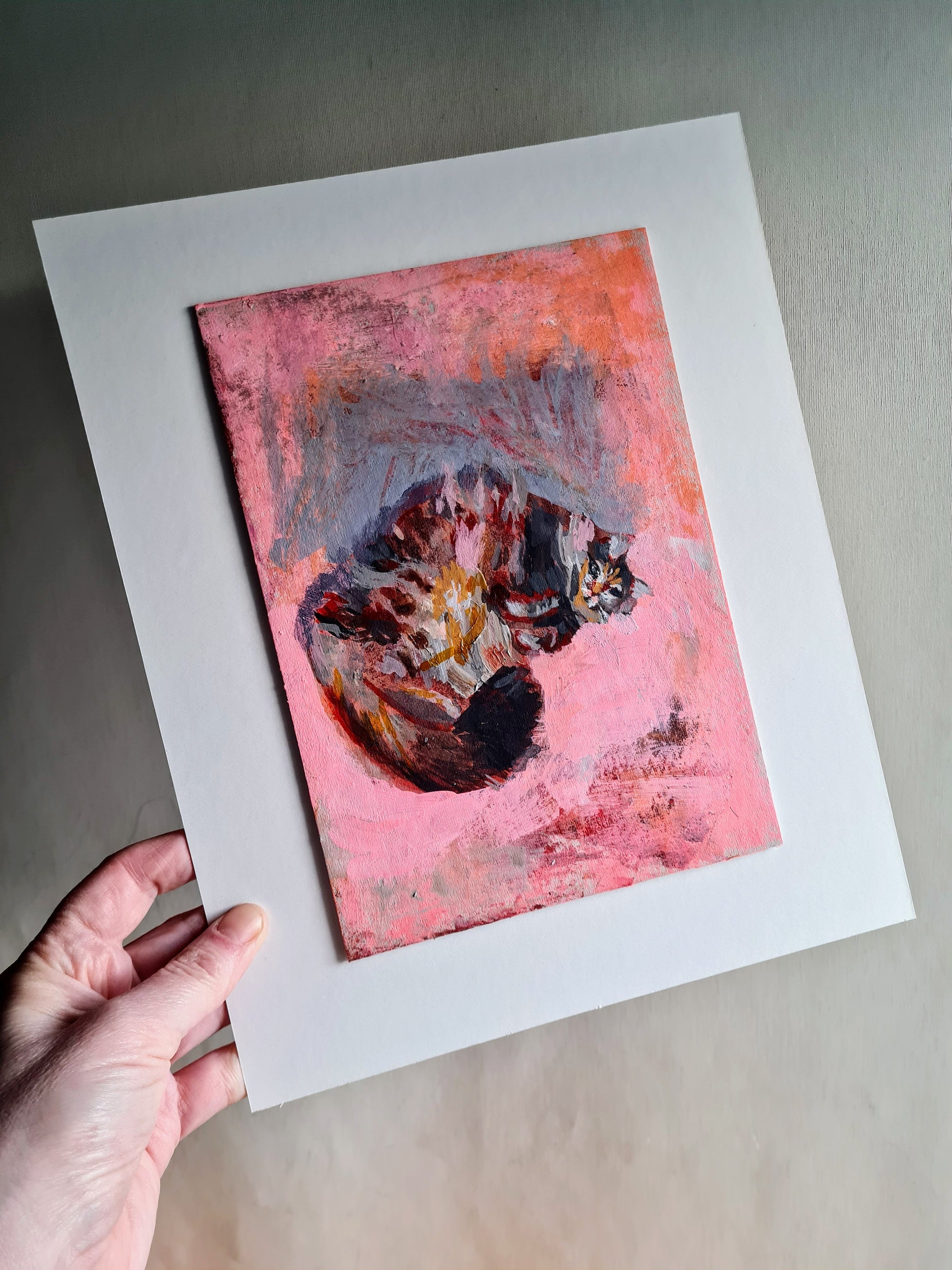 A white hand is holding up a small painting of a cat by Julia Laing.