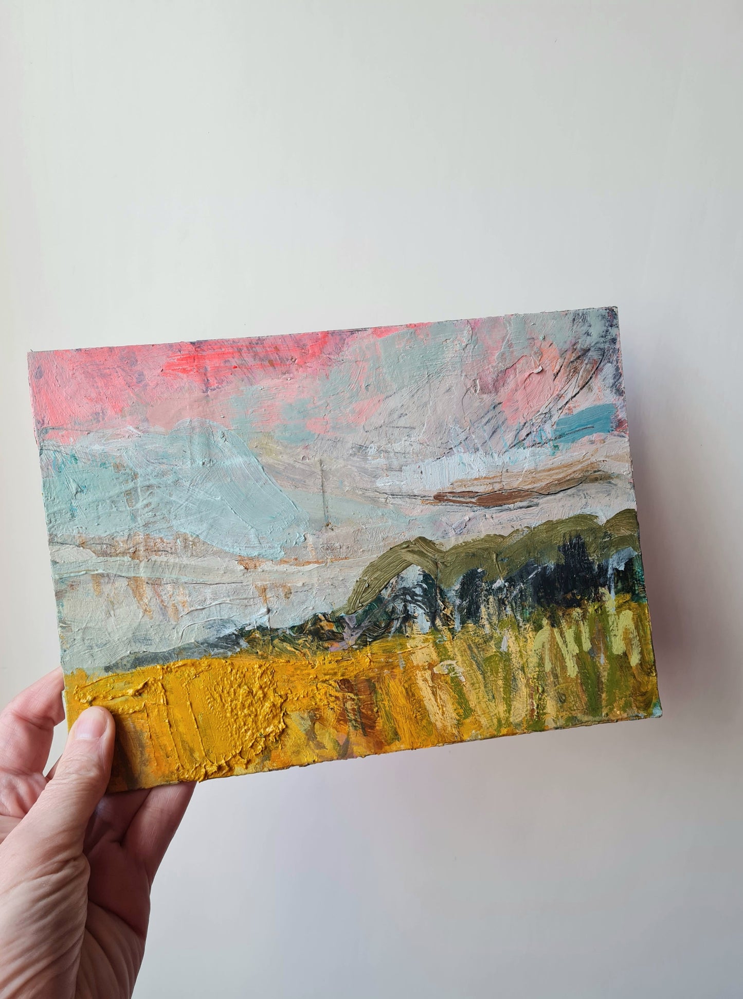 Hand holding a small abstract painting with a landscape scene on a white background