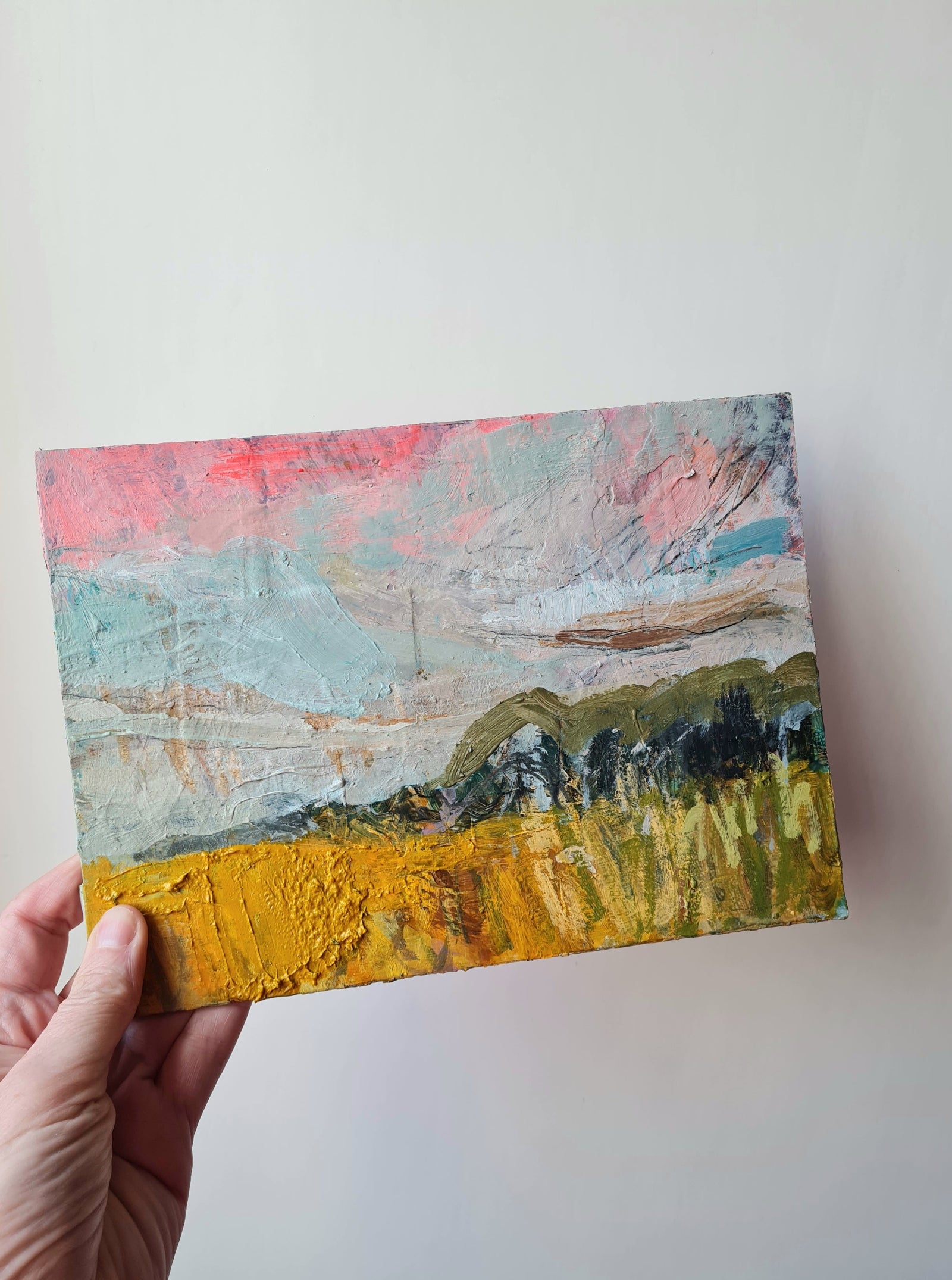 Hand holding a small abstract painting with a landscape scene on a white background
