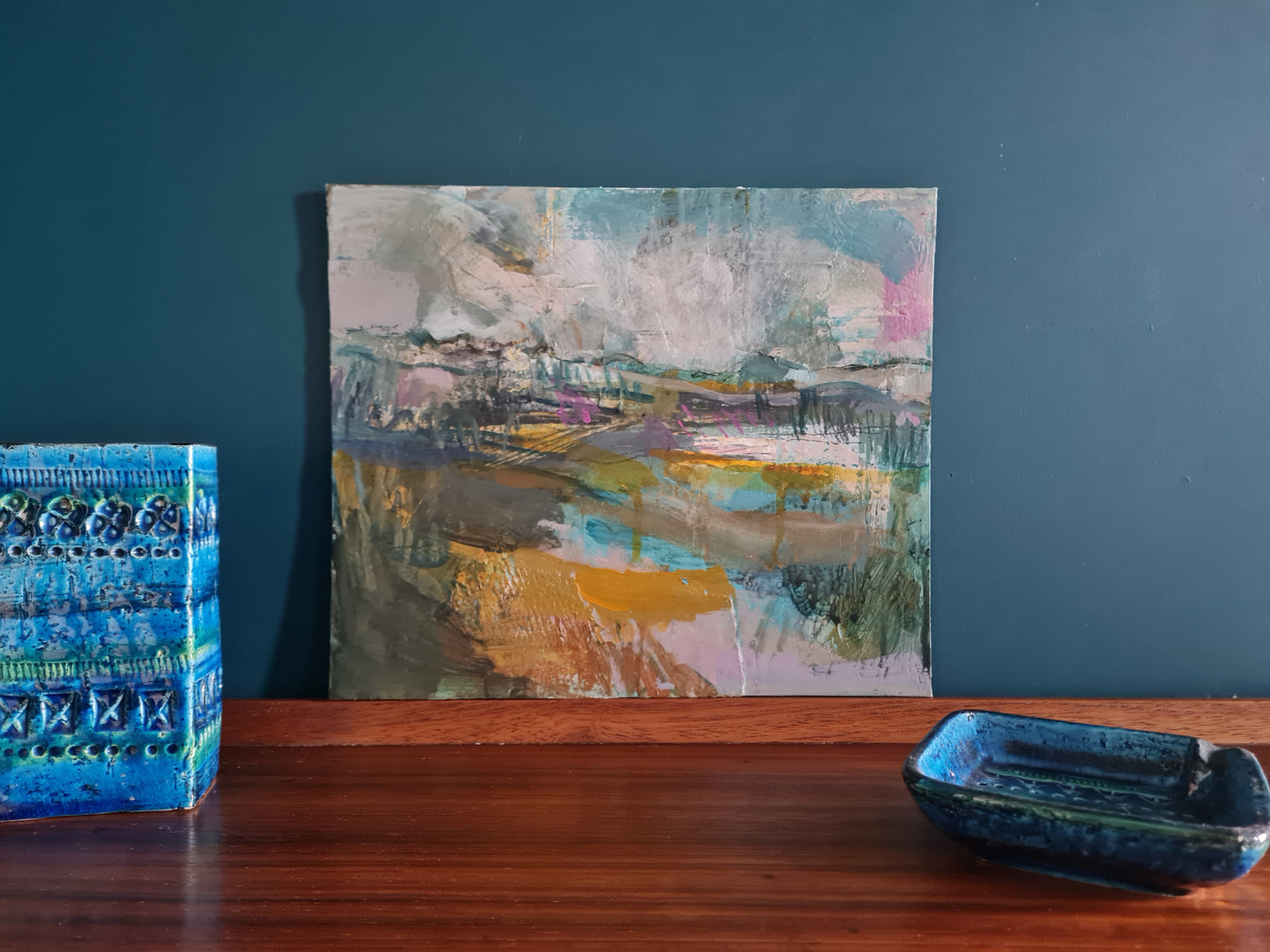 Abstract landscape painting on a dark wood surface with a blue rectangular hand built ceramic pot nearby.