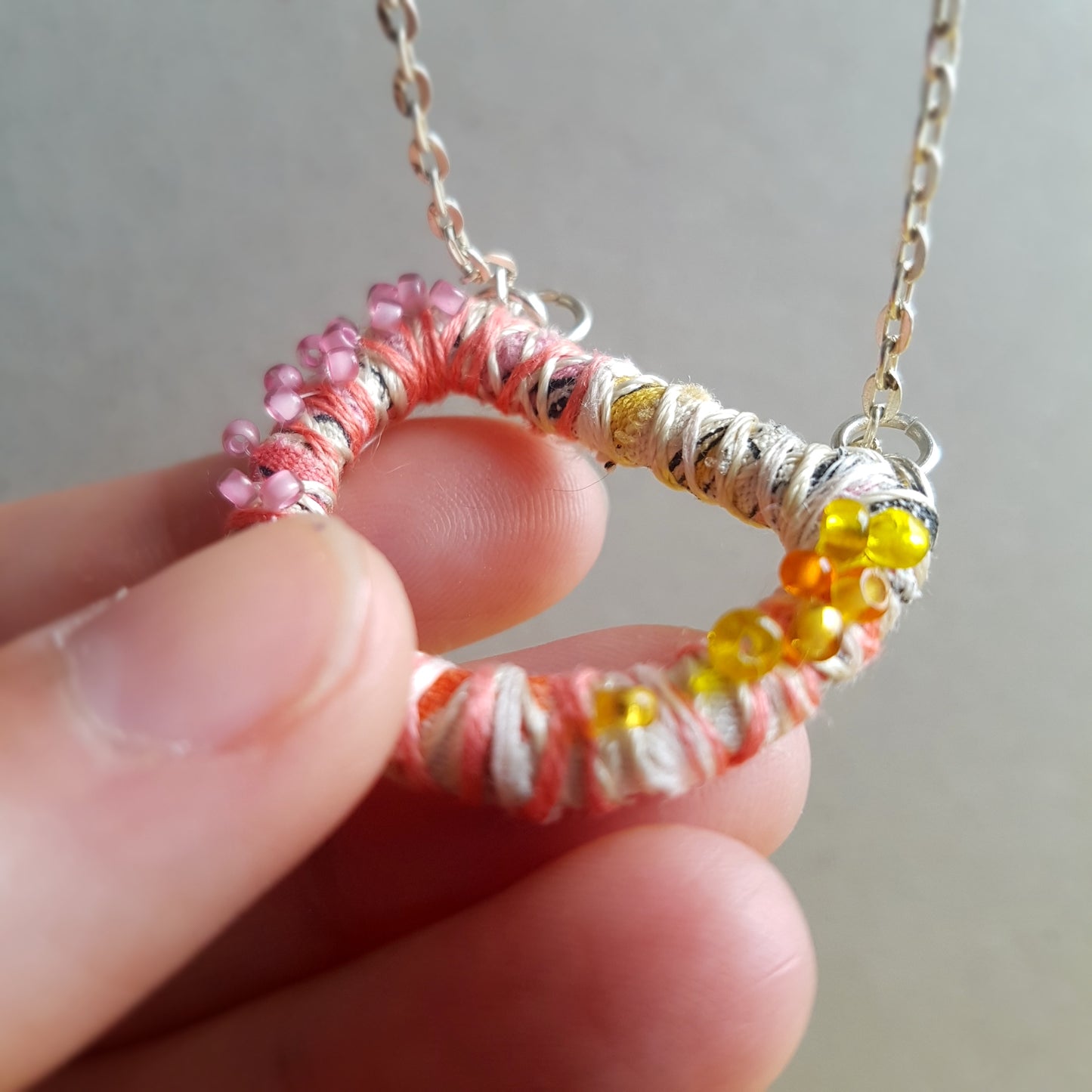 Pink cream and yellow textile art pendant necklace being held up to the camera by a white hand. Background is grey.