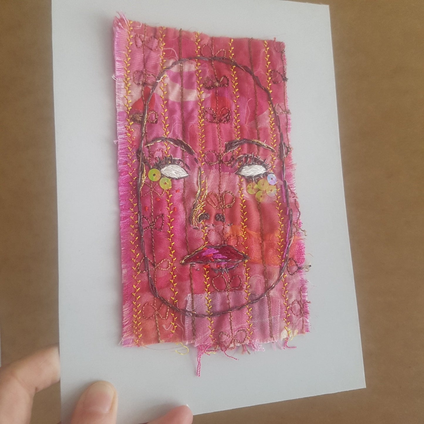 Textile-art embroidery of woman with blank eyes, mounted on backing board.