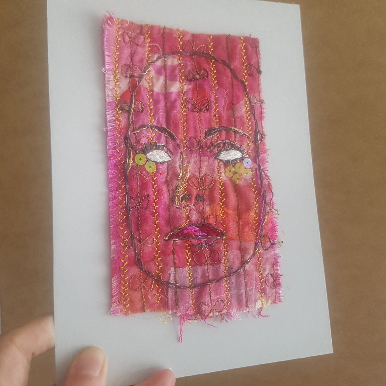 Textile-art embroidery of woman with blank eyes, mounted on backing board.
