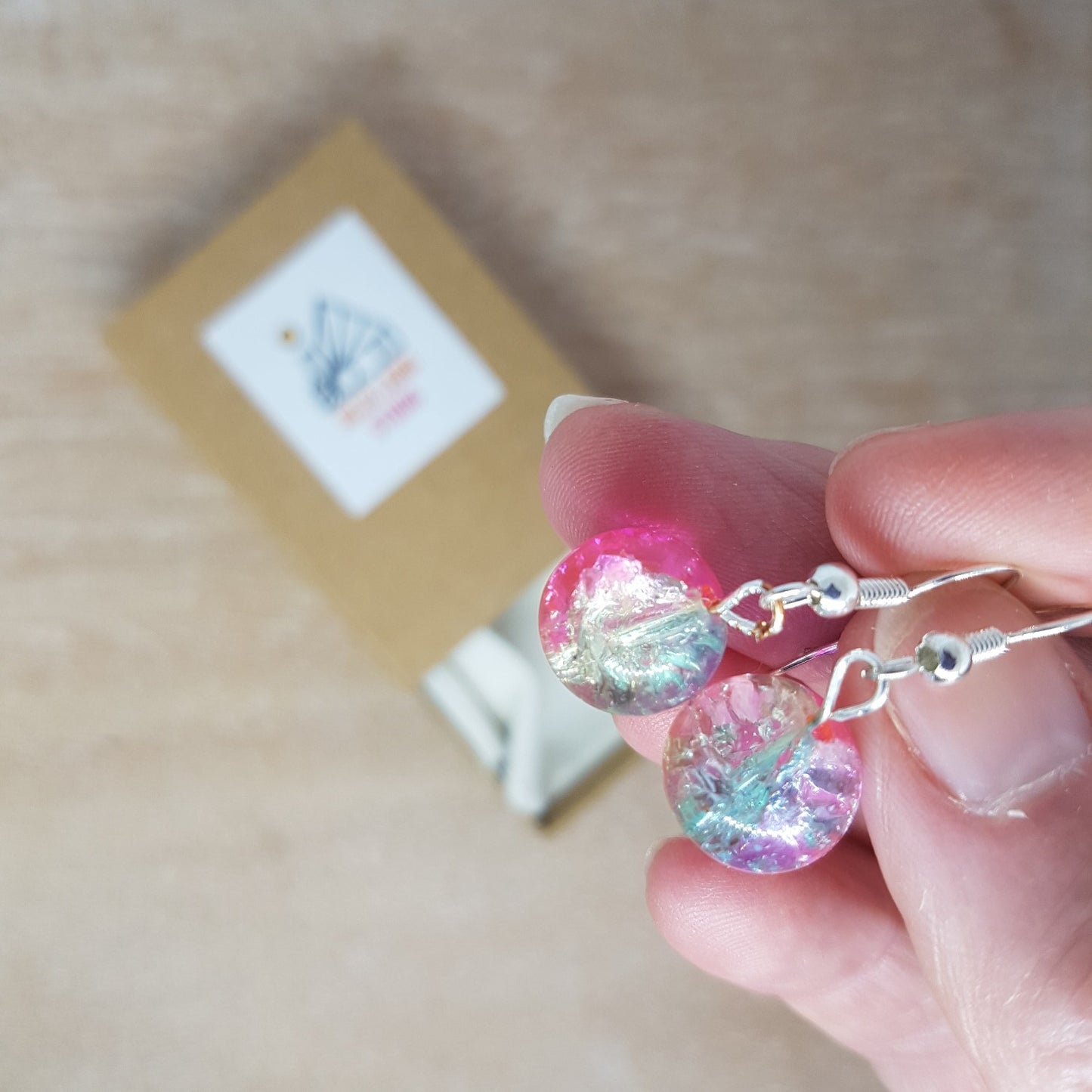 Hand holding pink sparkly glass bead drop earrings up to the camera. Brown craft card gift box can be seen on the desk below.