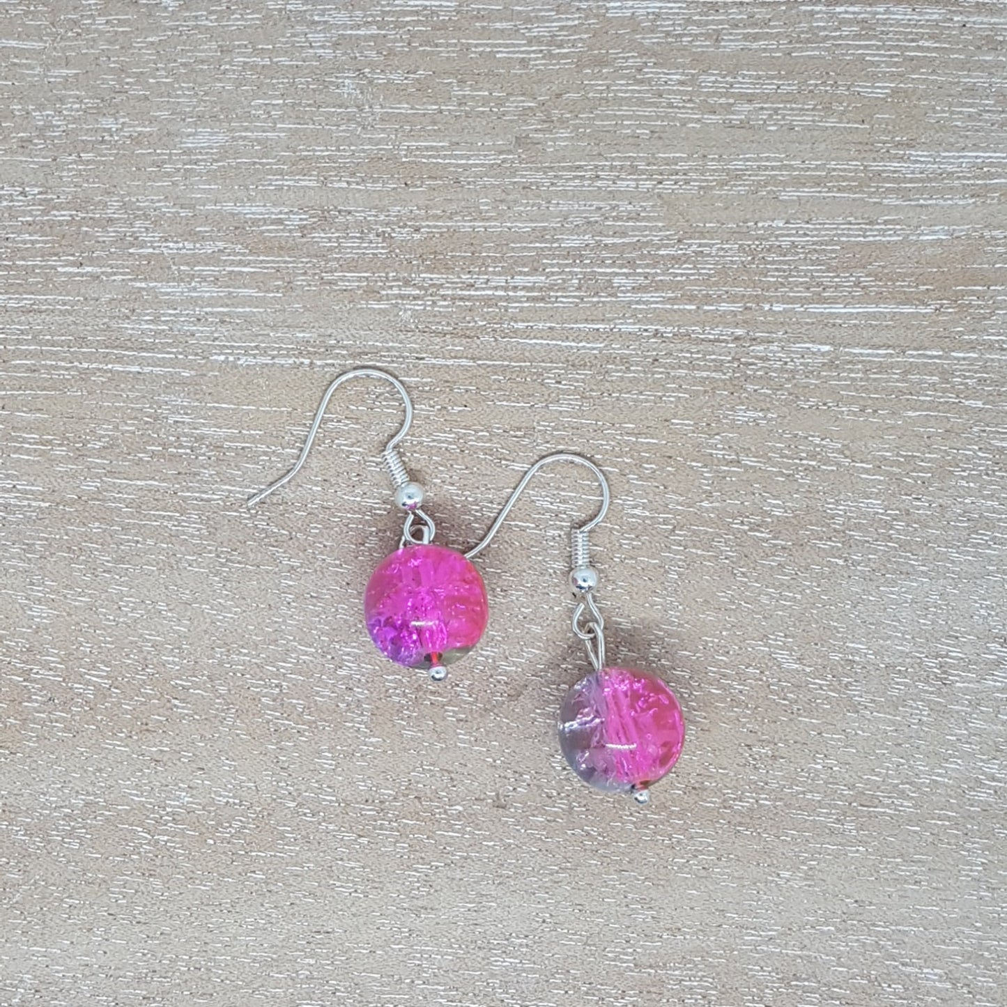 A pair of silver plated drop earrings with pink sparkly beads, sitting on a wooden table .