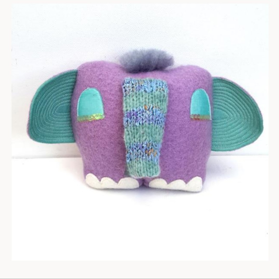 Tufty is a cute elephant soft sculpture doll made by Julia Laing from lilac wool with big blue quilted silk ears. He is sitting on a white table.