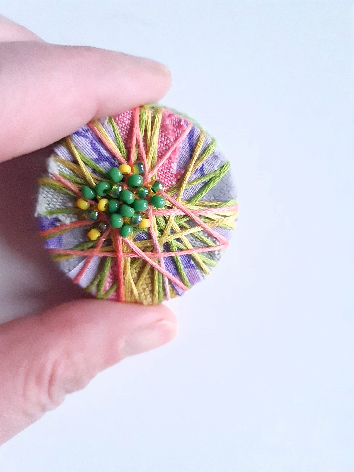 colourful textile art statement brooch held between fingers against a light background