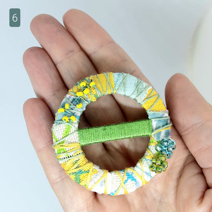 Hand holding a statement textile brooch which is yellow blue and green. Background is white.