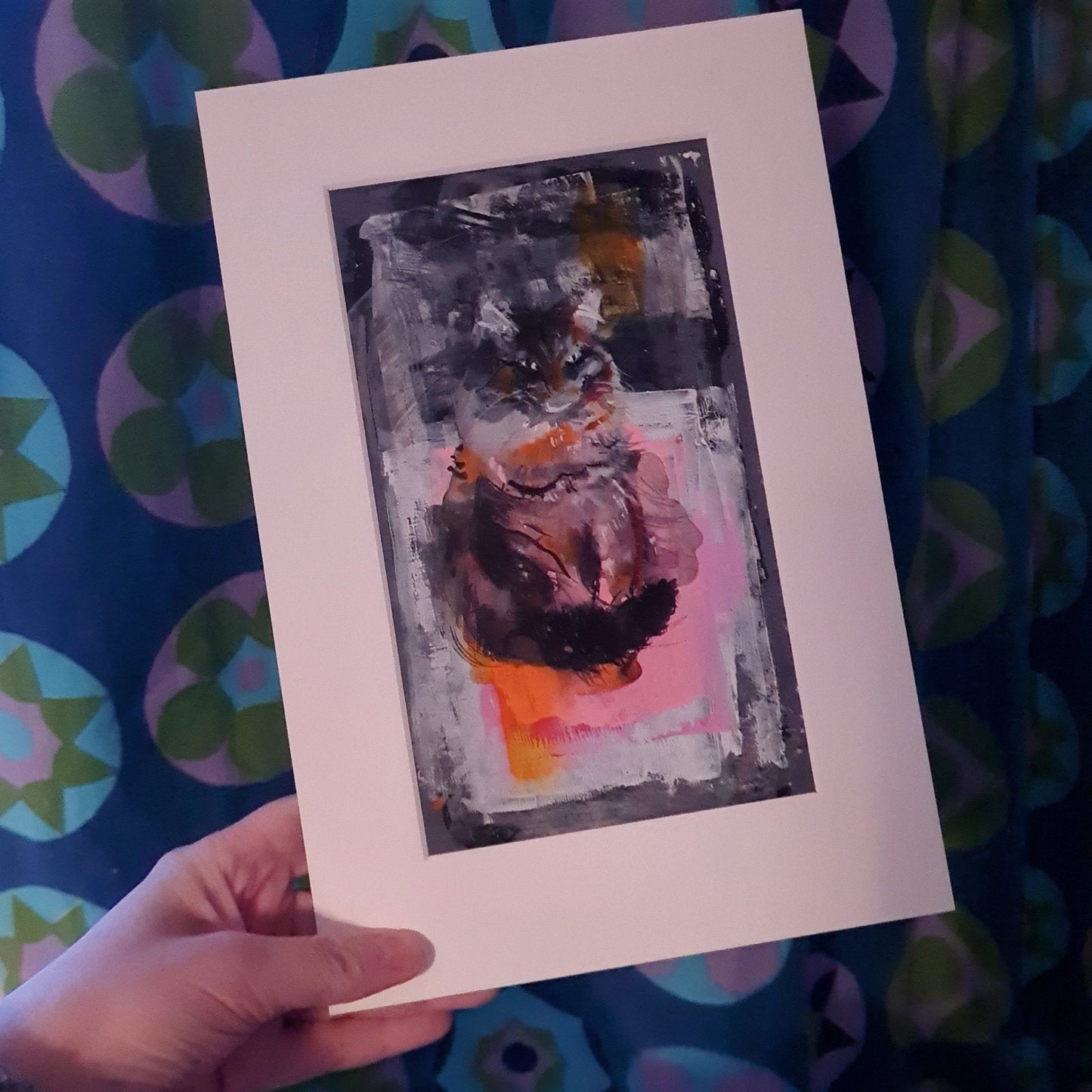 A hand holds up a small expressive acrylic painting of a cat. The painting is mounted with an ivory coloured window mount. There's a 1960s blue geometric patterned curtain in the background.