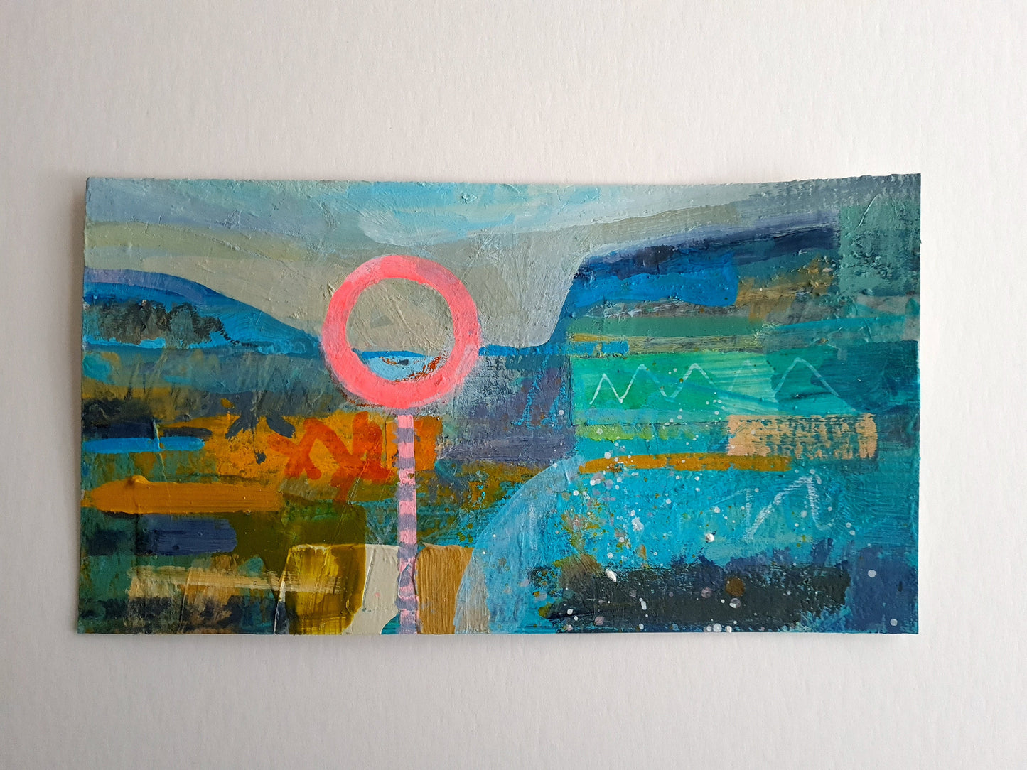 Colourful abstract painting with a neon pink life ring in the foreground and colourful blue and mustard landscape in the background, placed on a white table.
