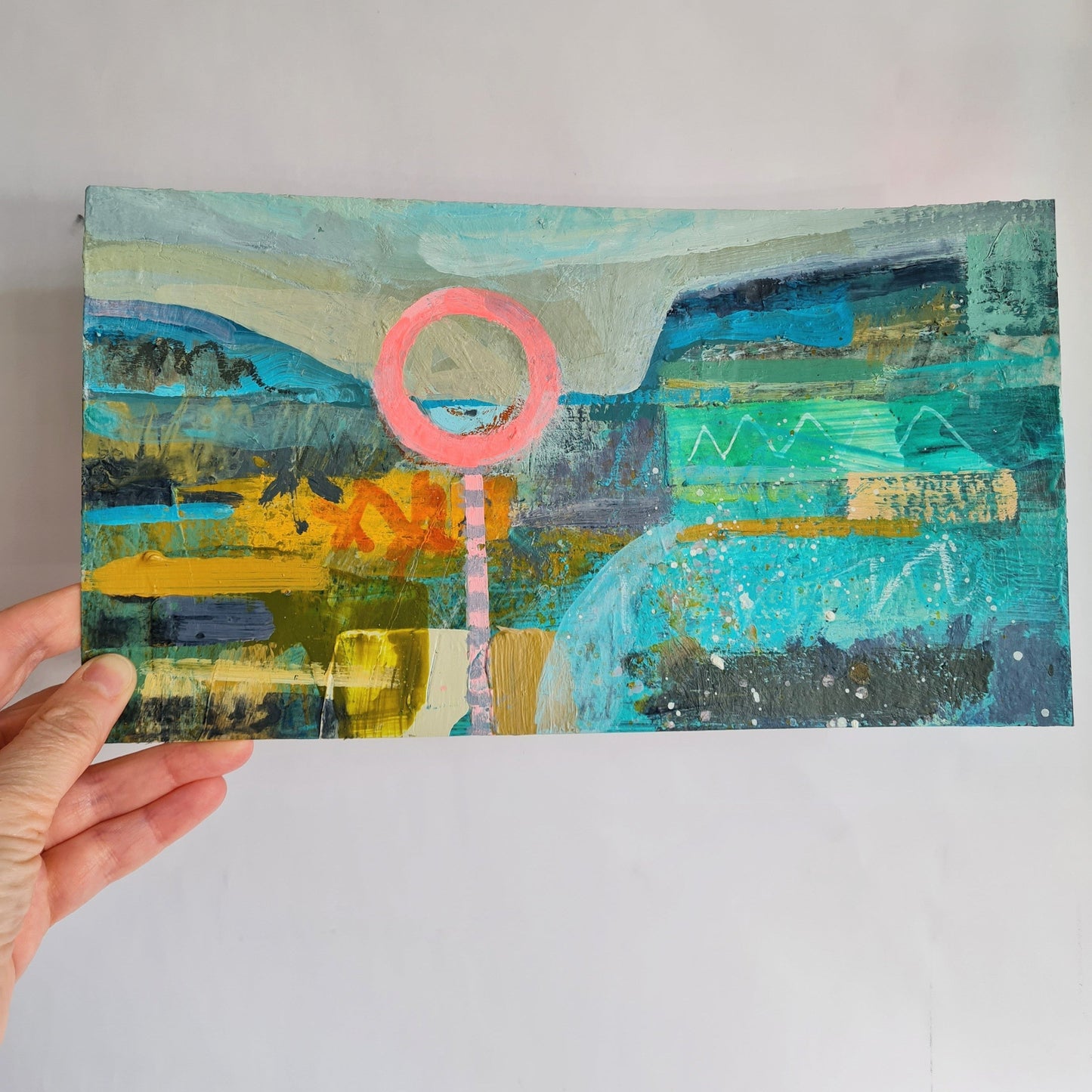 Hand holding a colourful abstract painting with a pink circle in the foreground and blue, mustard and grey colours in the background. The painting is held up against a white background
