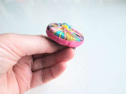 Hand holding a colourful brooch with a pink fabric cover against a light background