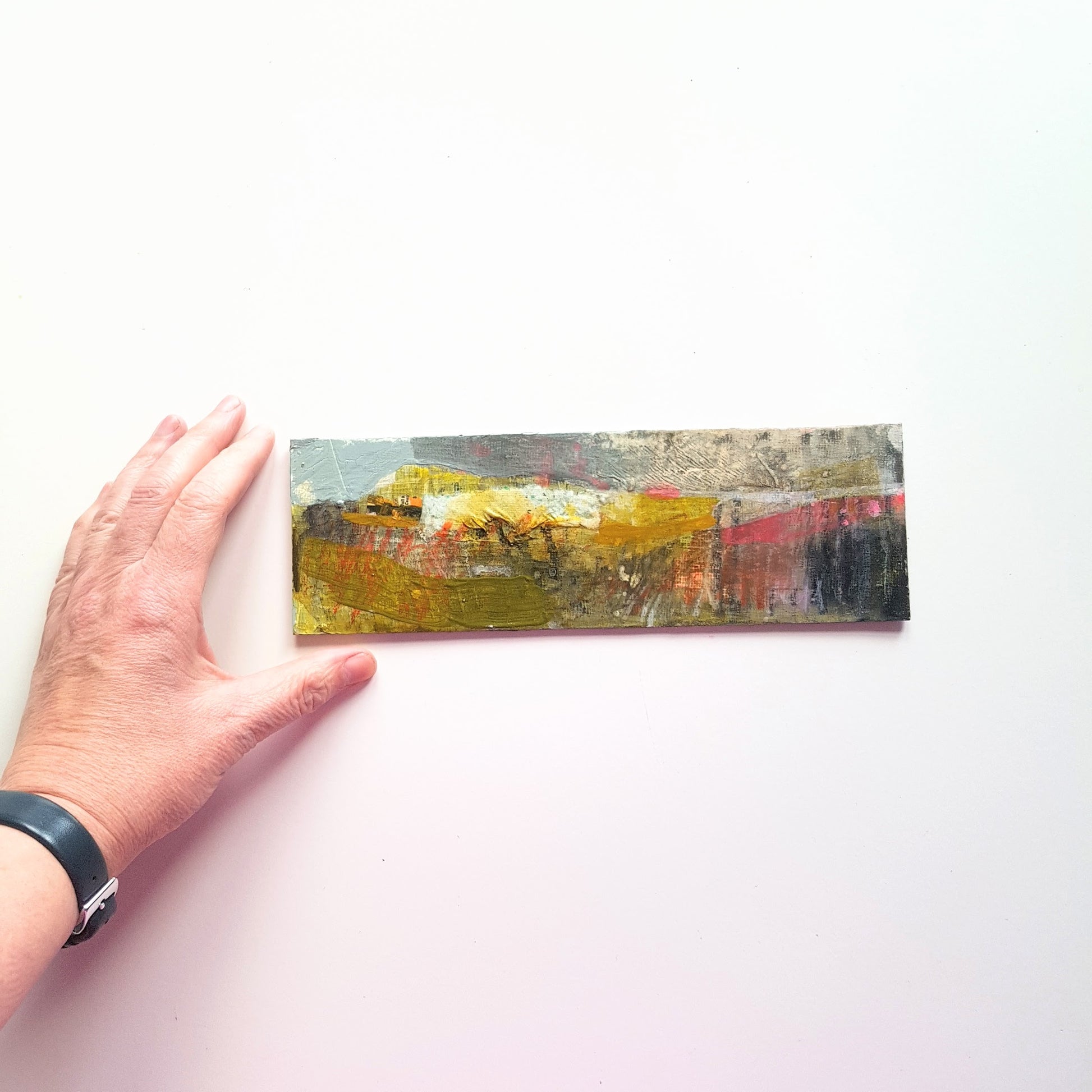 small horizontal mixed media landscape on a white table with a hand beside it for scale.