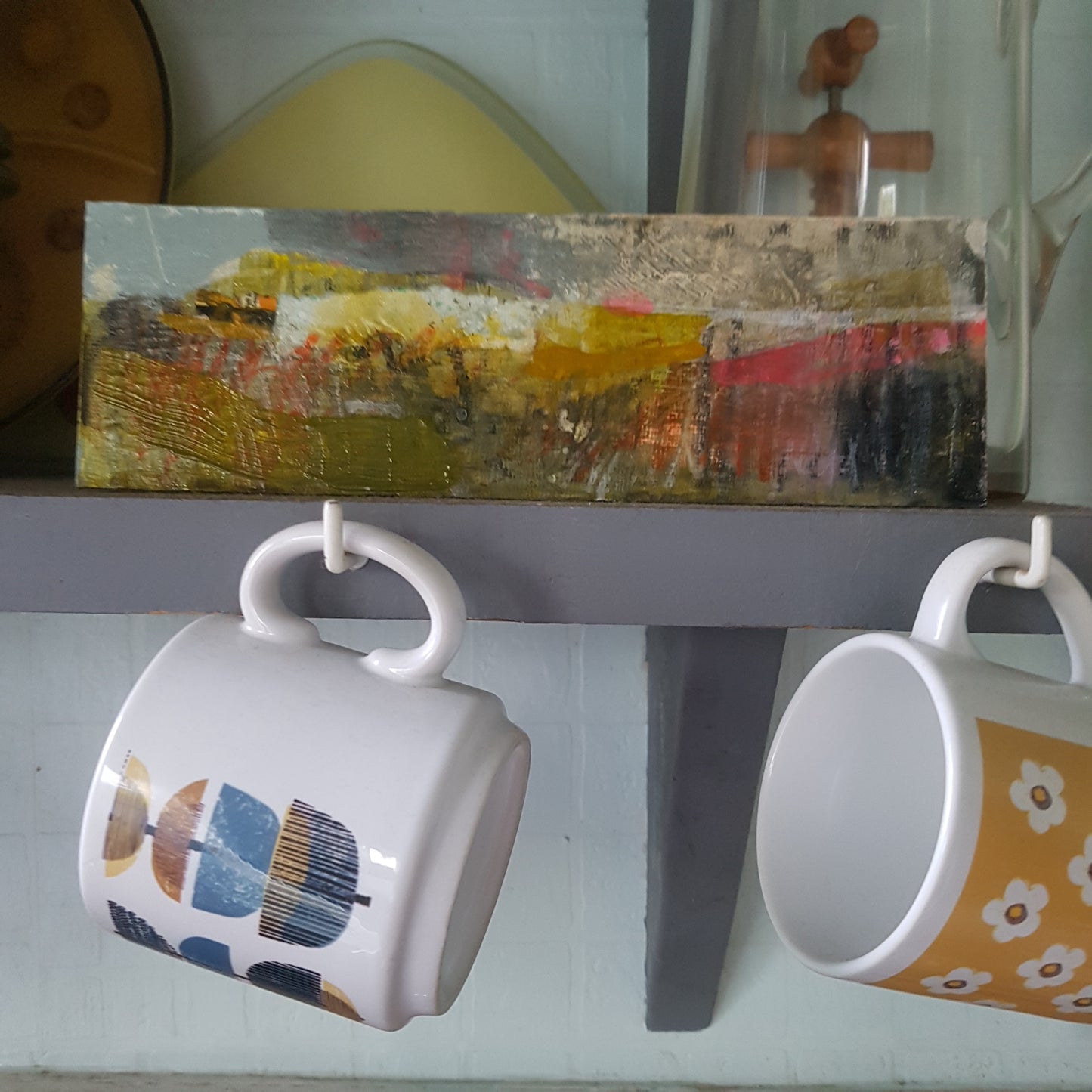 Small mixed-media landscape painting on akitchen shelf.