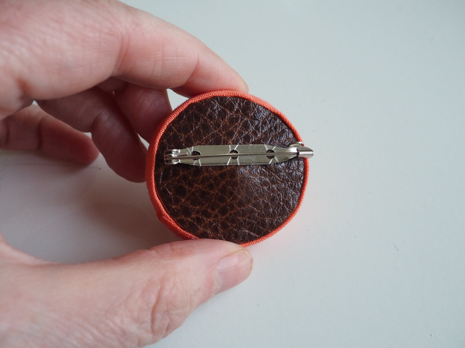 Back of brooch made of brown leather with a metal fastening pin, against a white background