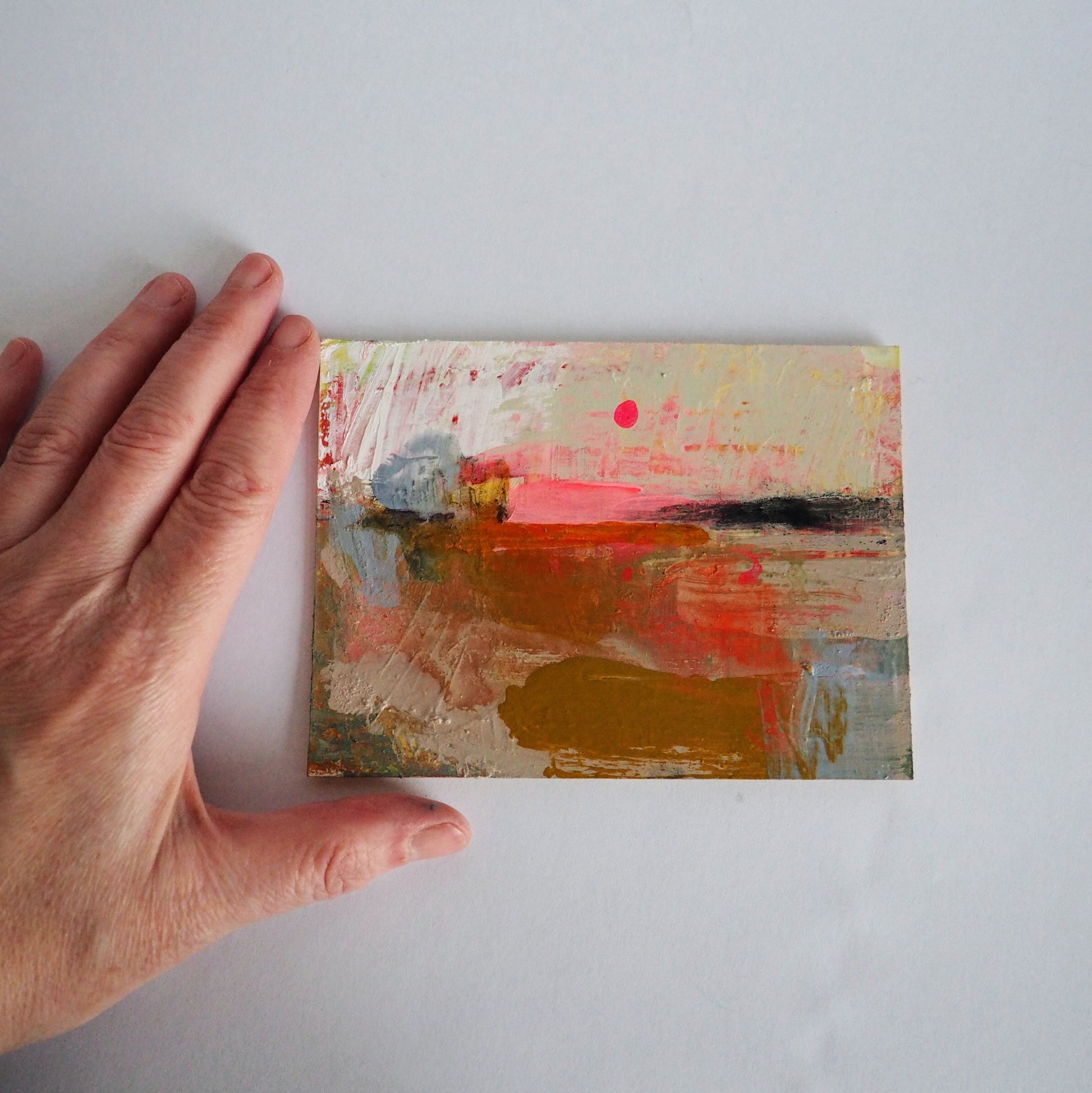 Mini expressive landscape painting on a white desk, with a hand beside it to show scale.