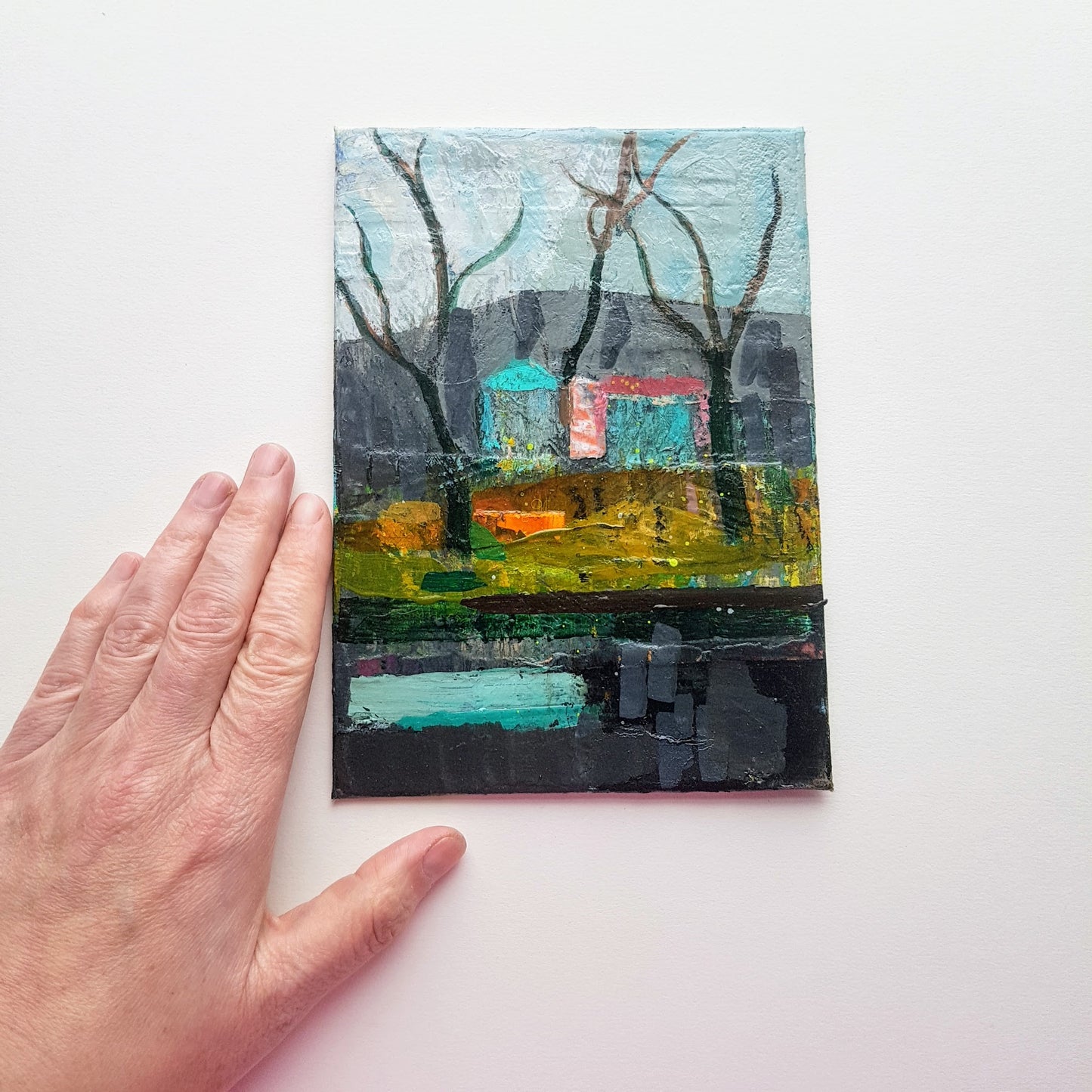 Small painted scene, Winter Walk by Scottish artist Julia Laing, lying on a white table with a hand for scale.