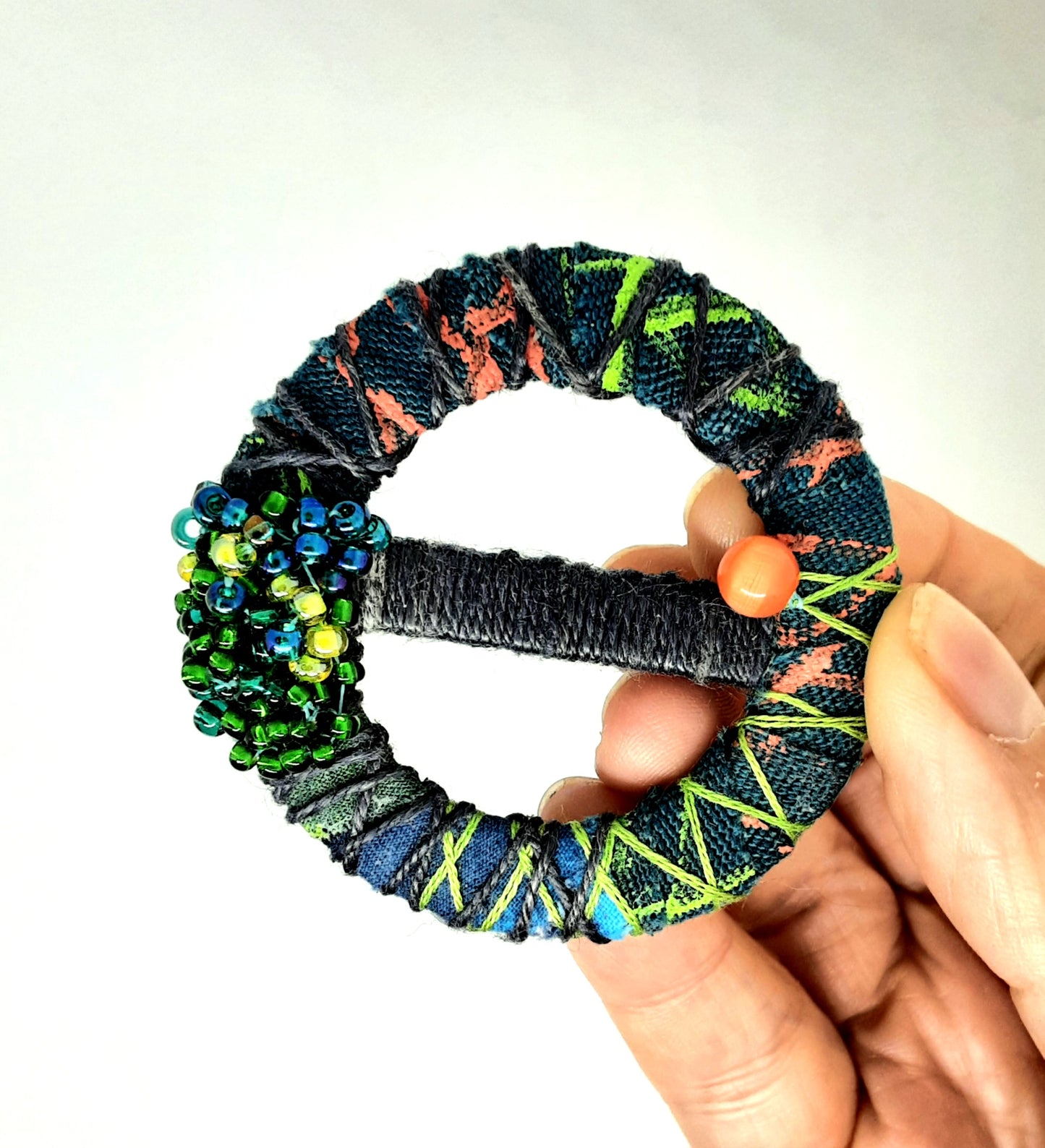 Hand holding up a fabric wrapped and beaded textile brooch. Background is white.