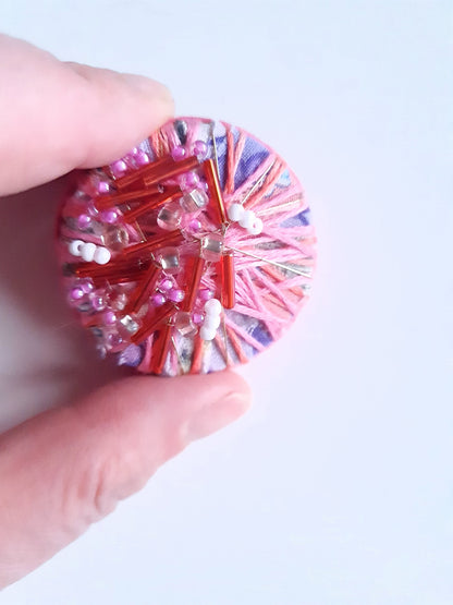 Fingers holding textile art brooch against a white background.