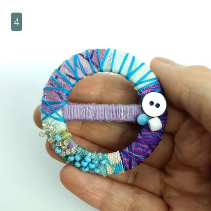 Blue and purple statement brooch with bead ad button detail being held up to the camera. Background is white.