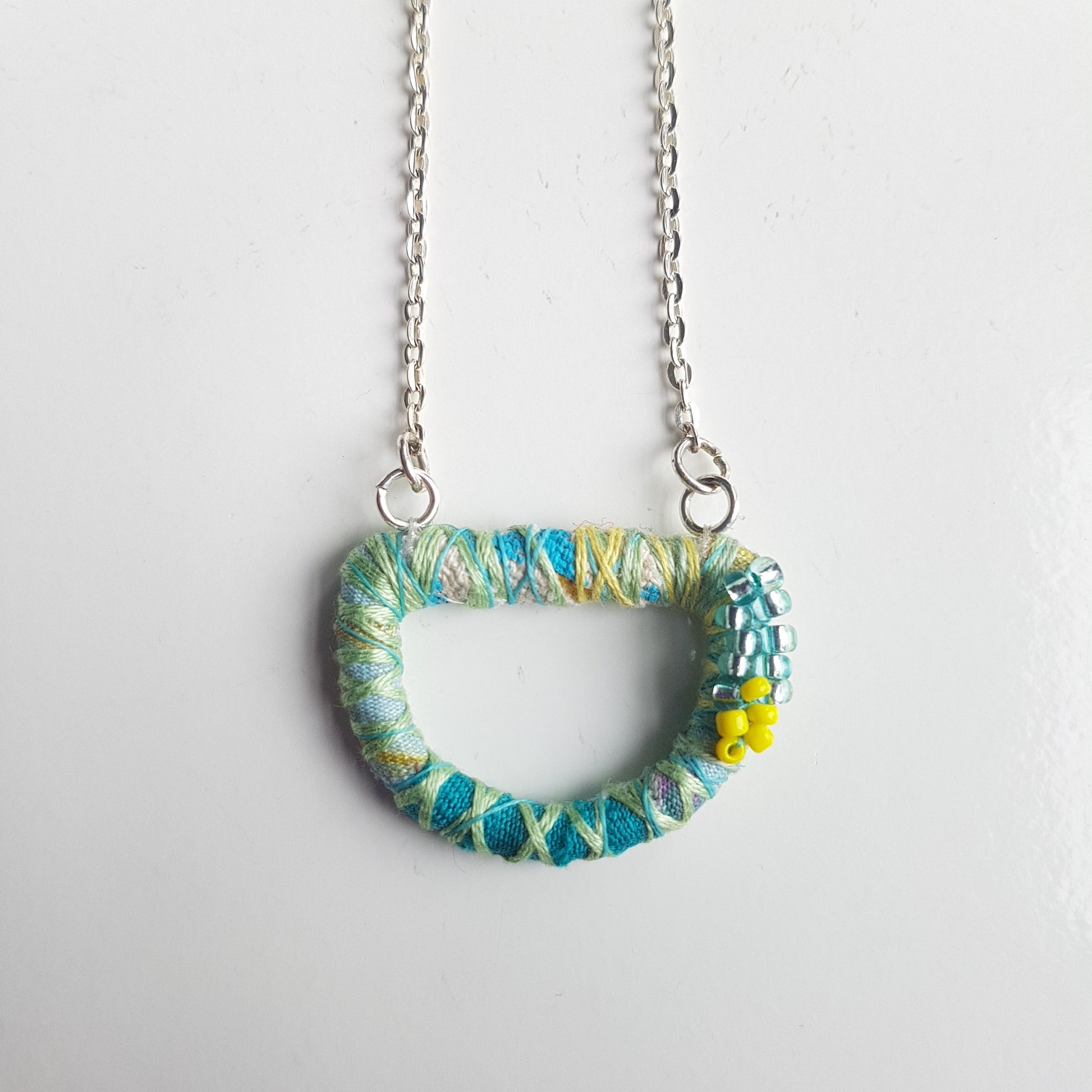 Pale green  blue and yellow textile art necklace with an 18" silver plated chain on a white table top.