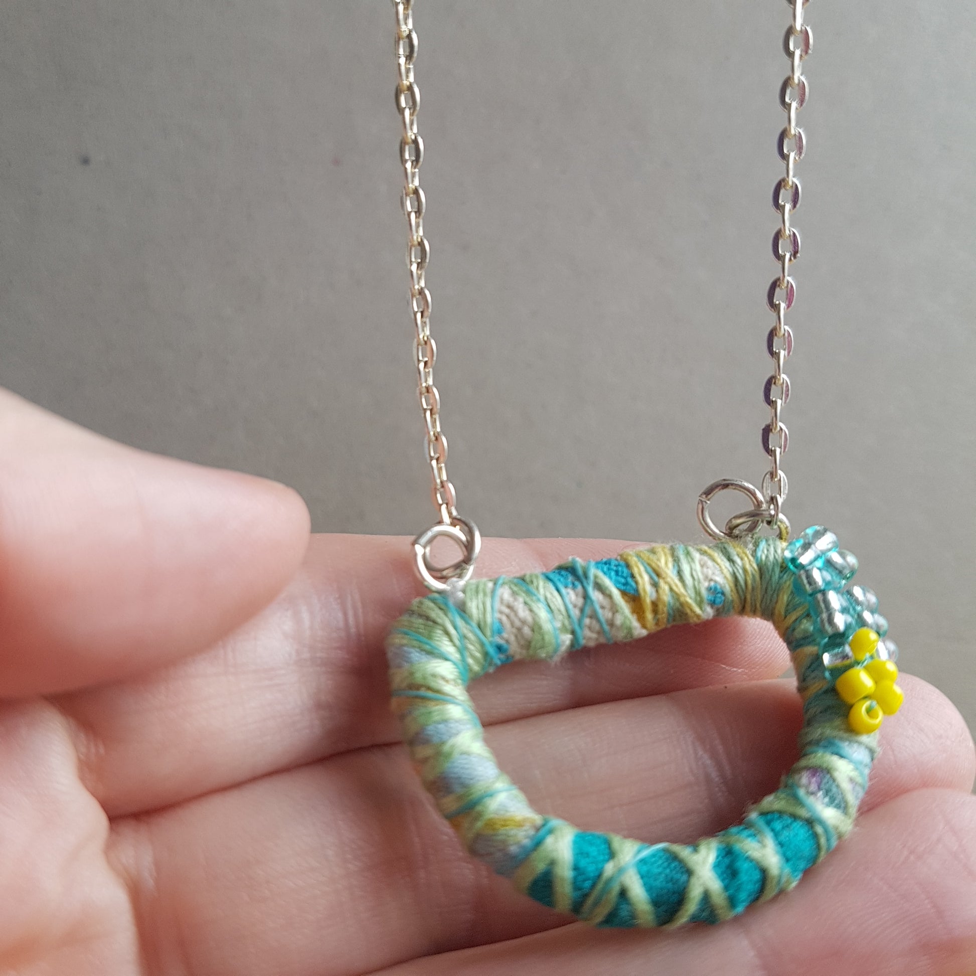 Detail of textile art necklace being held up the the camera by a white hand. There are lemon yellow seed bead embellishments and green threads wrapped around a D ring shaped pendant.