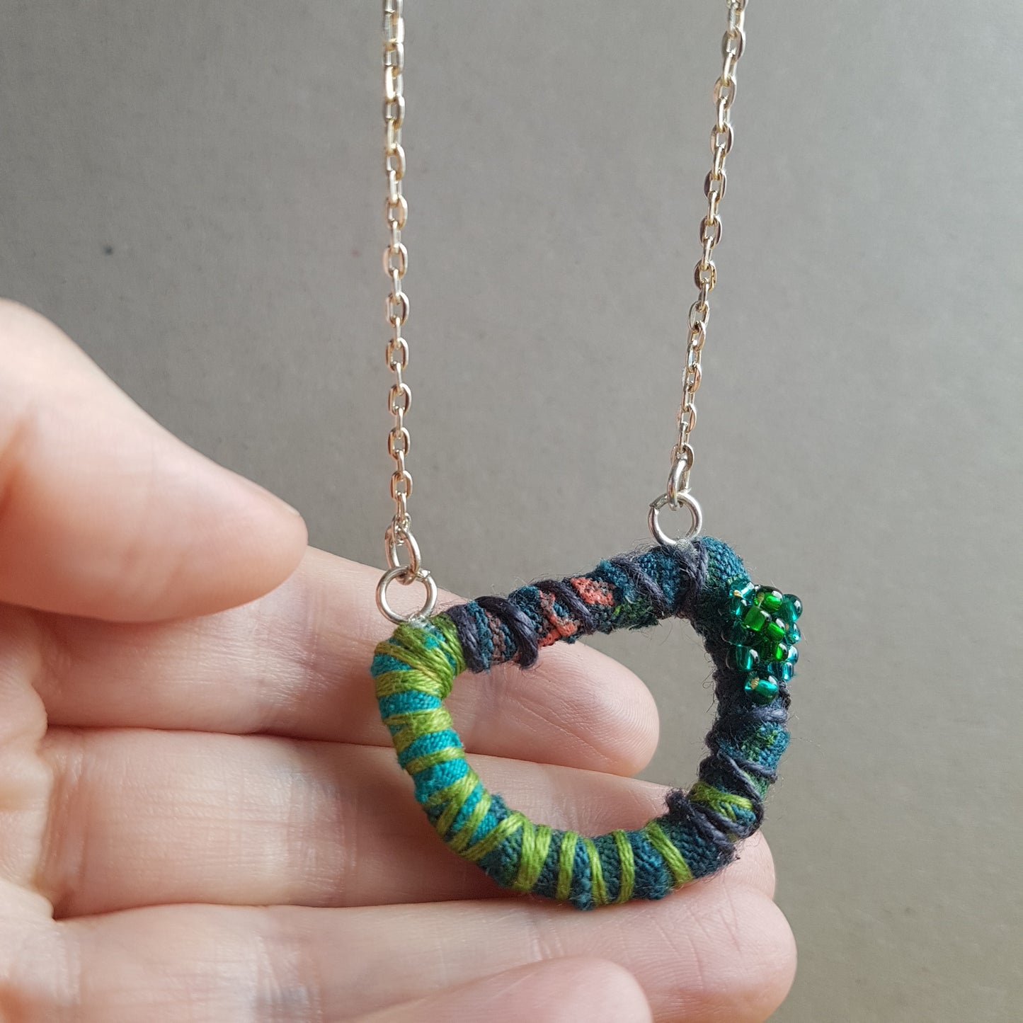 Hand holding up a teal and green textile art necklace to the camera. The background is grey.