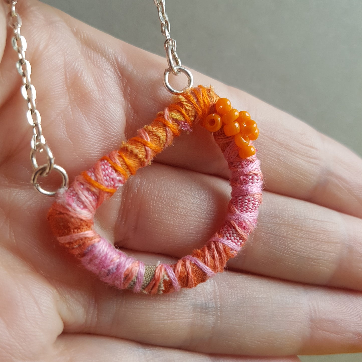 Hand holding a pink and orange textile art necklace with a silver plated chain.