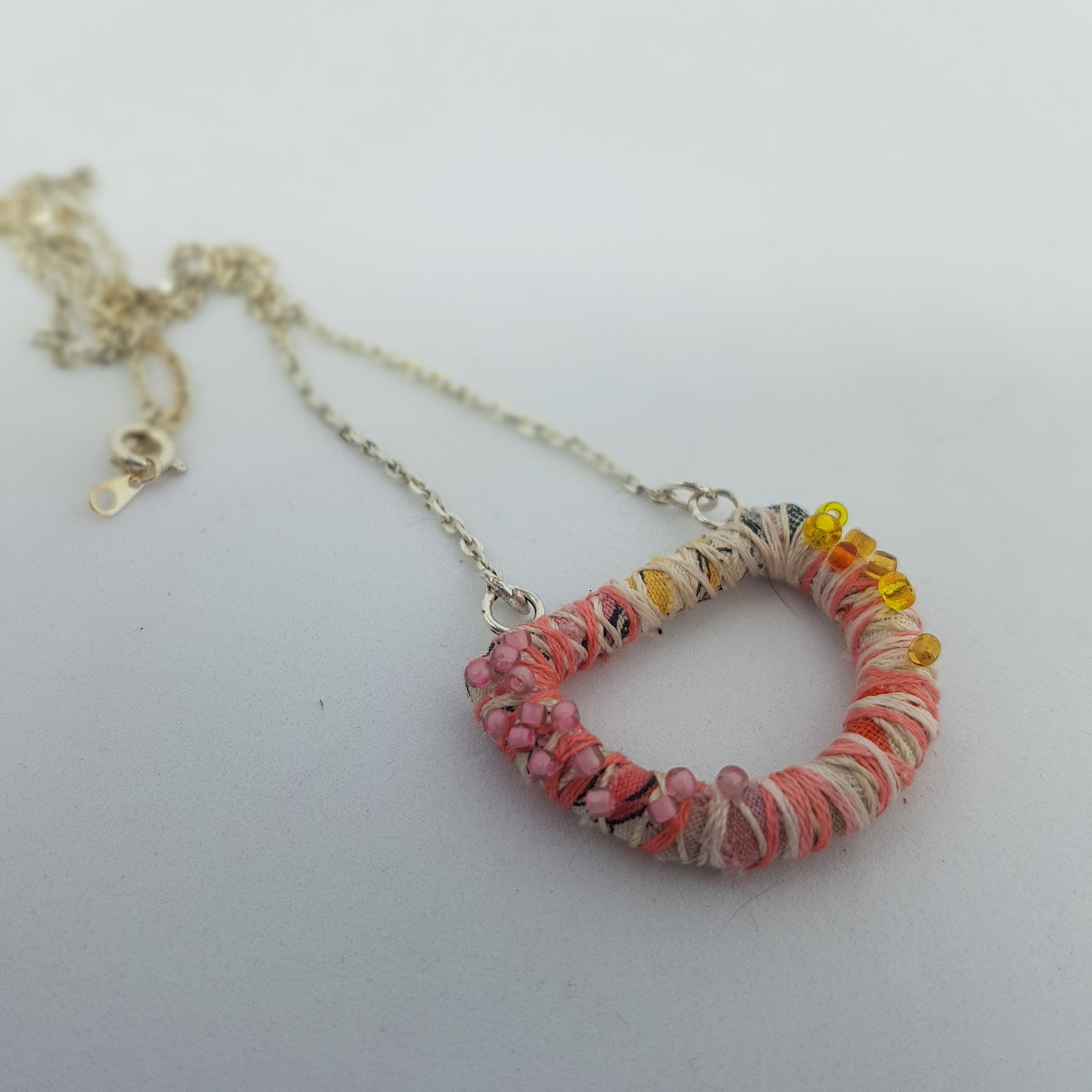 Pink and cream D-ring shaped textile art necklace , with seed bead detail lying on a white table top.