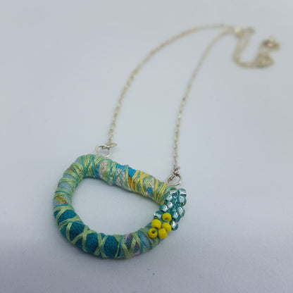 Blue and yellow textile art necklace with seed bead embellishments lying on a white table.