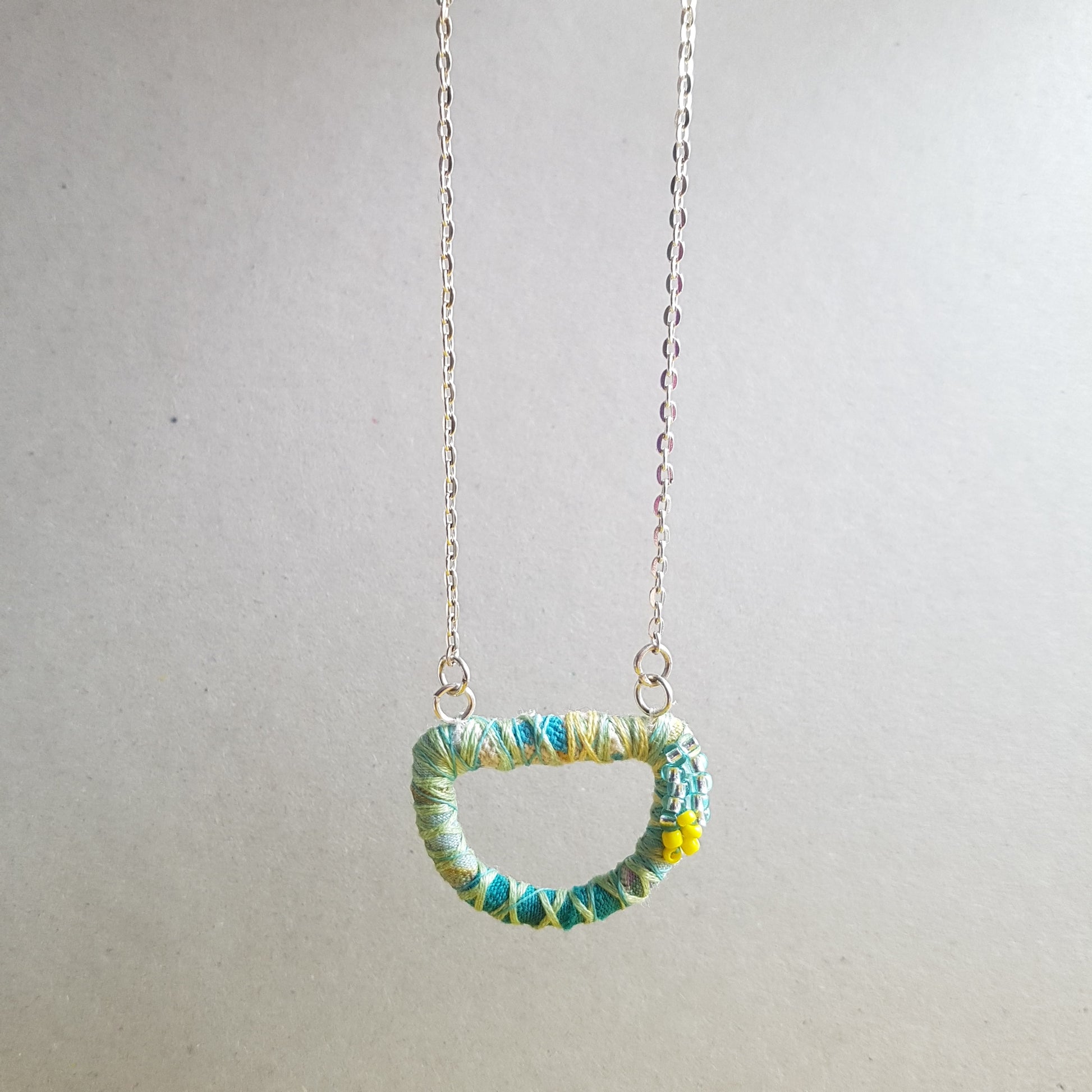 Textile art necklace with 18" silver plated chain hanging against a grey background.
