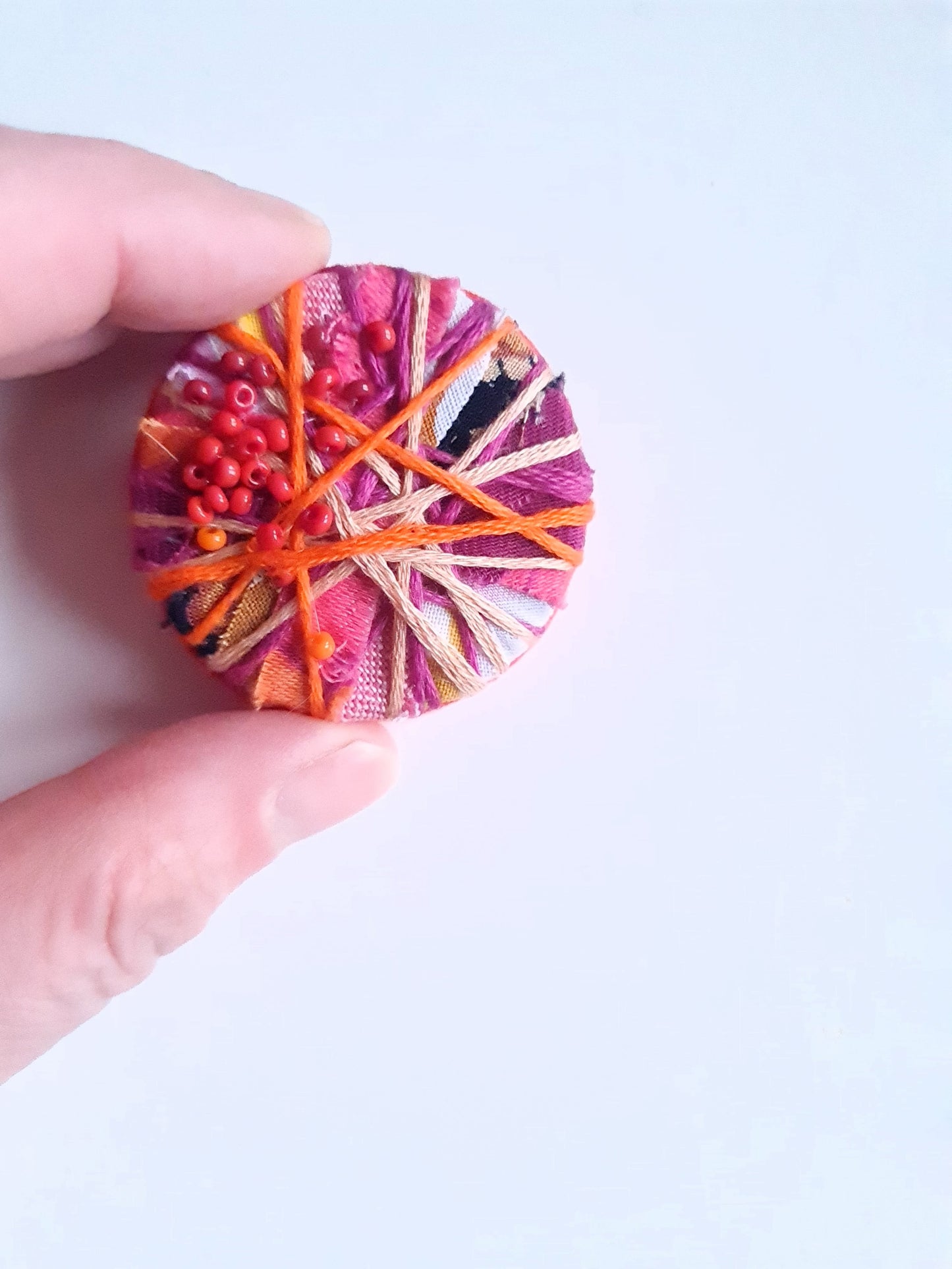 Hand holding a colorful fabric brooch with yarn and beads on a light background