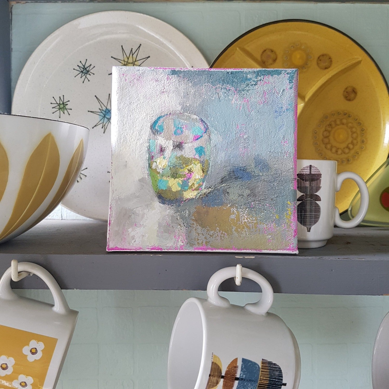Painting by Julia Laing sitting on a shelf beside midcentury design bowls and plates.