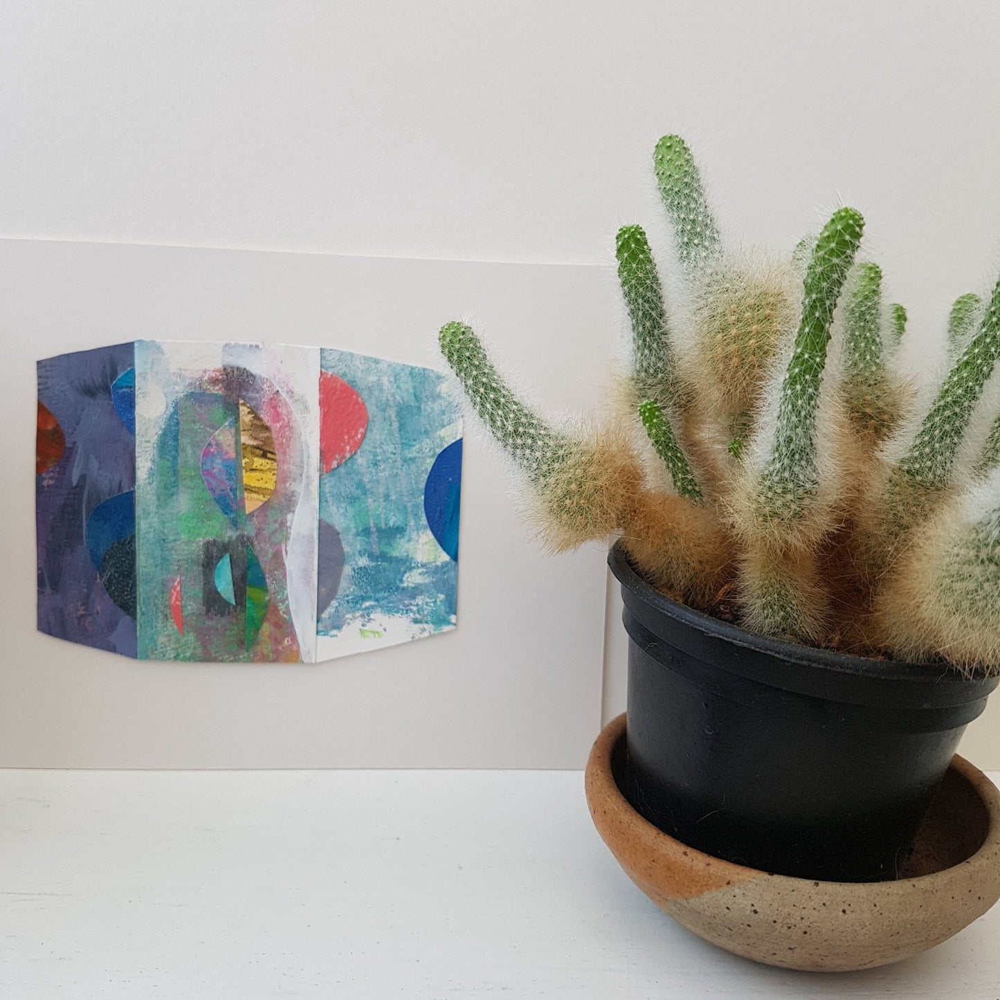 Mixed media paper collage on a shelf beside a potted cactus.