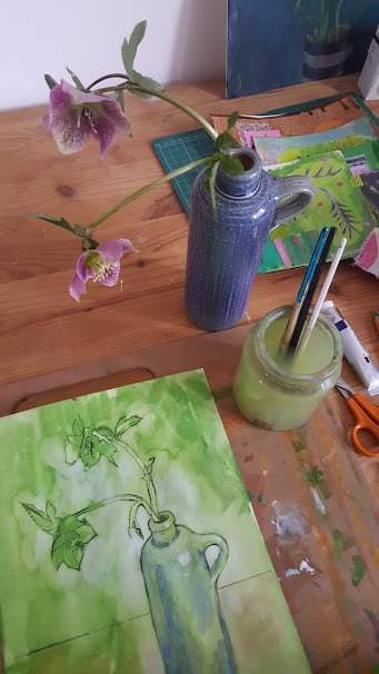 Painting in progress. Hellebore flowers in a ceramic bottle on a desk, beside the painting in progress.