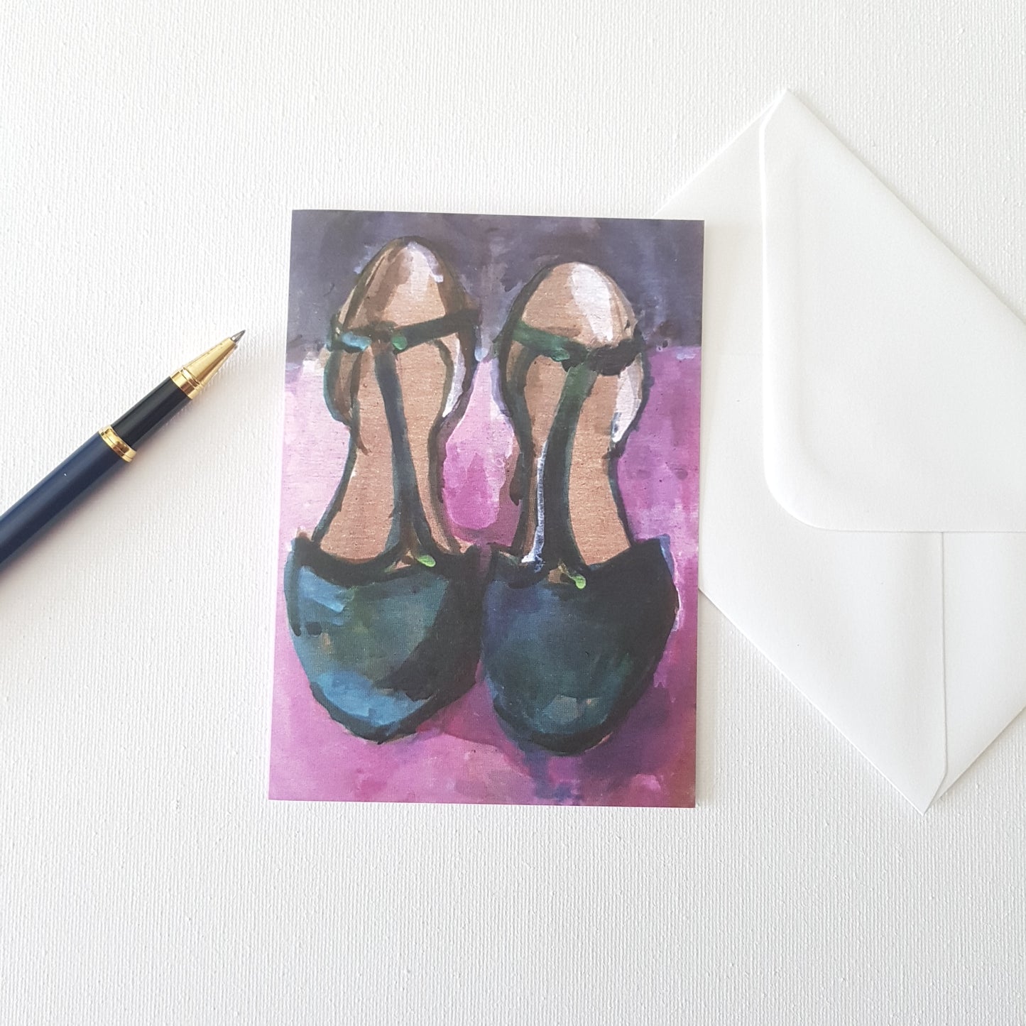 Shoe art greeting card featuring a painting of green t-bar shoes on a pink background by Julia Laing. Placed beside the card on a white table are a pen and white envelope.