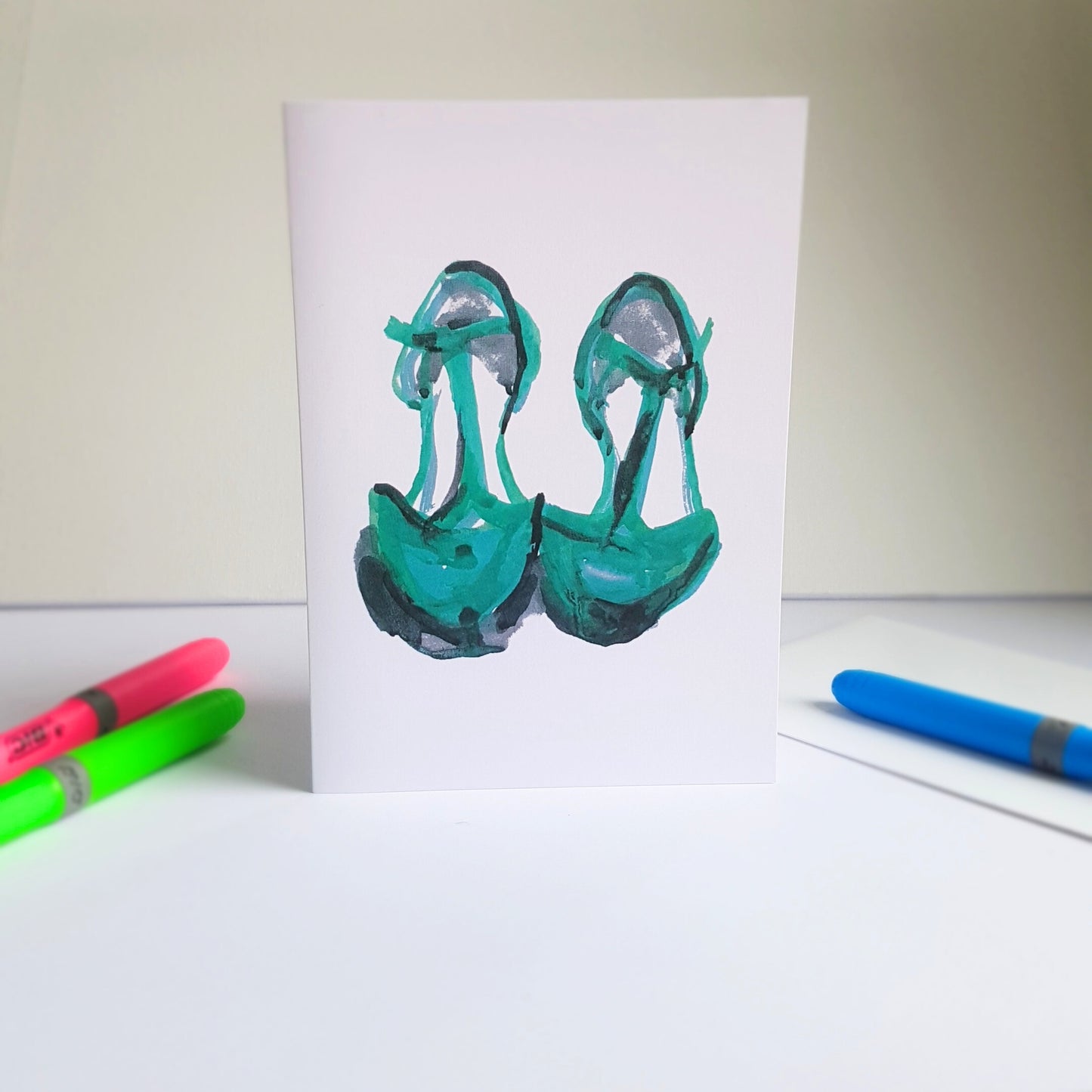 Shoe art greeting card featuring an illustration of green t-bar shoes by UK artist Julia Laing. Beside the card are  neon marker pens and a white envelope.