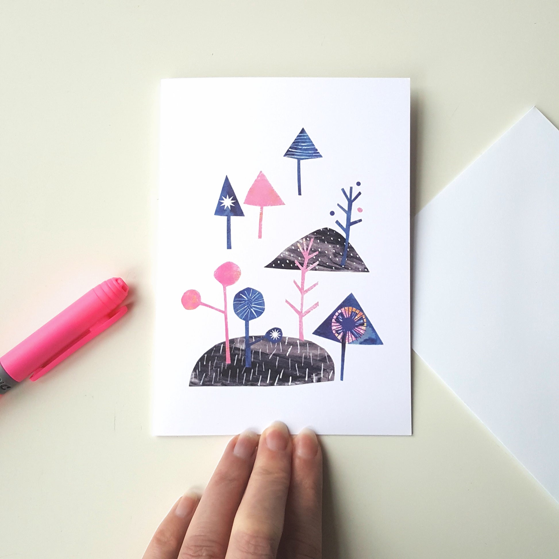 Whimsical Woods greeting card with a hand holding it down on a white table with a white envelope and neon pink pen lying beside it.